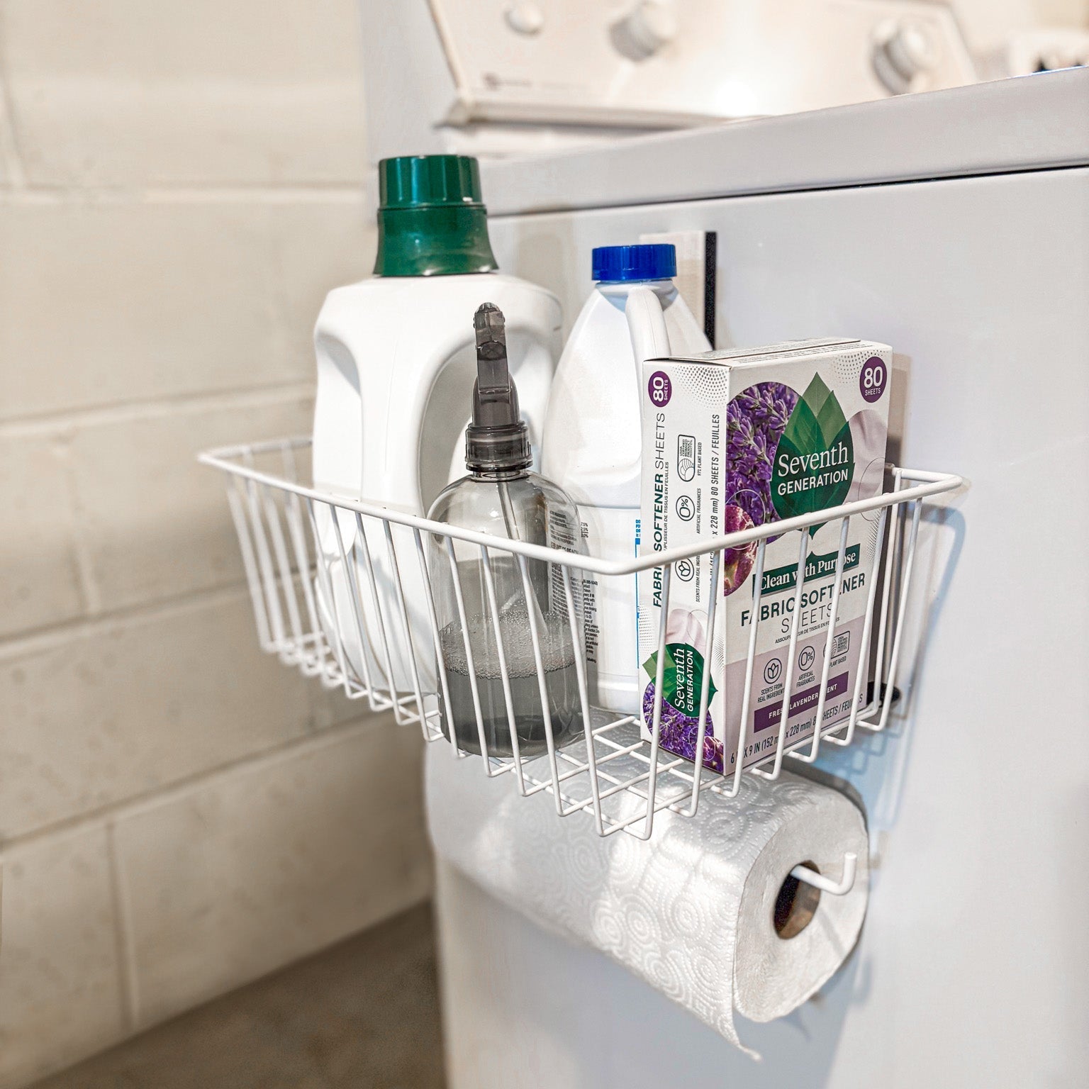 A white magnetic storage basket from Gator Magnetics holds laundry detergent, fabric softener, spray bottle, and fabric softener sheets, attached to the side of a white steel washing machine with a paper towel roll hanging below using a gator magnetic hook.