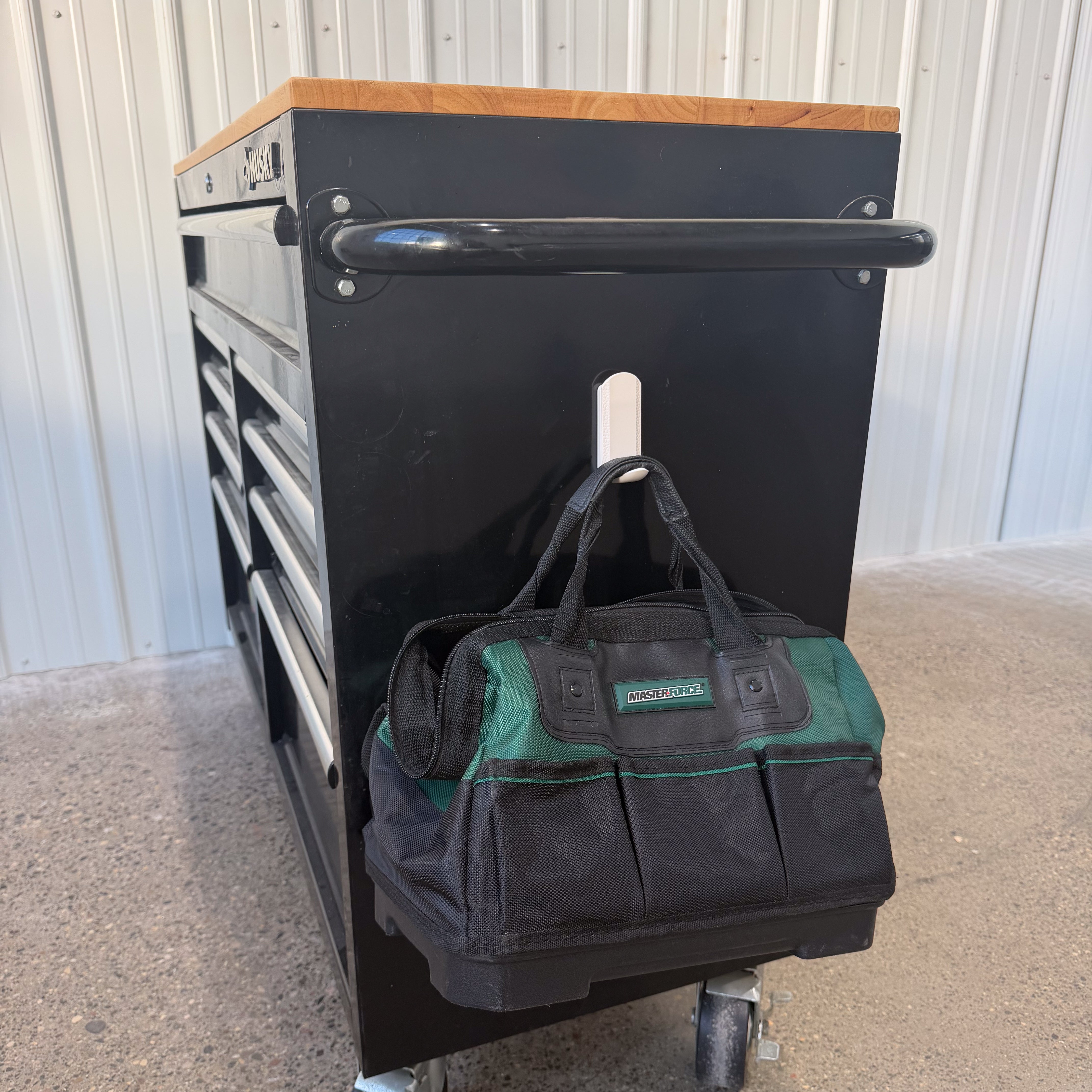 A green and black tool bag is hanging from a white magnetic hook attached to the steel side panel of a black tool chest in a workshop, demonstrating practical hook tools storage with Gator Magnetics products.