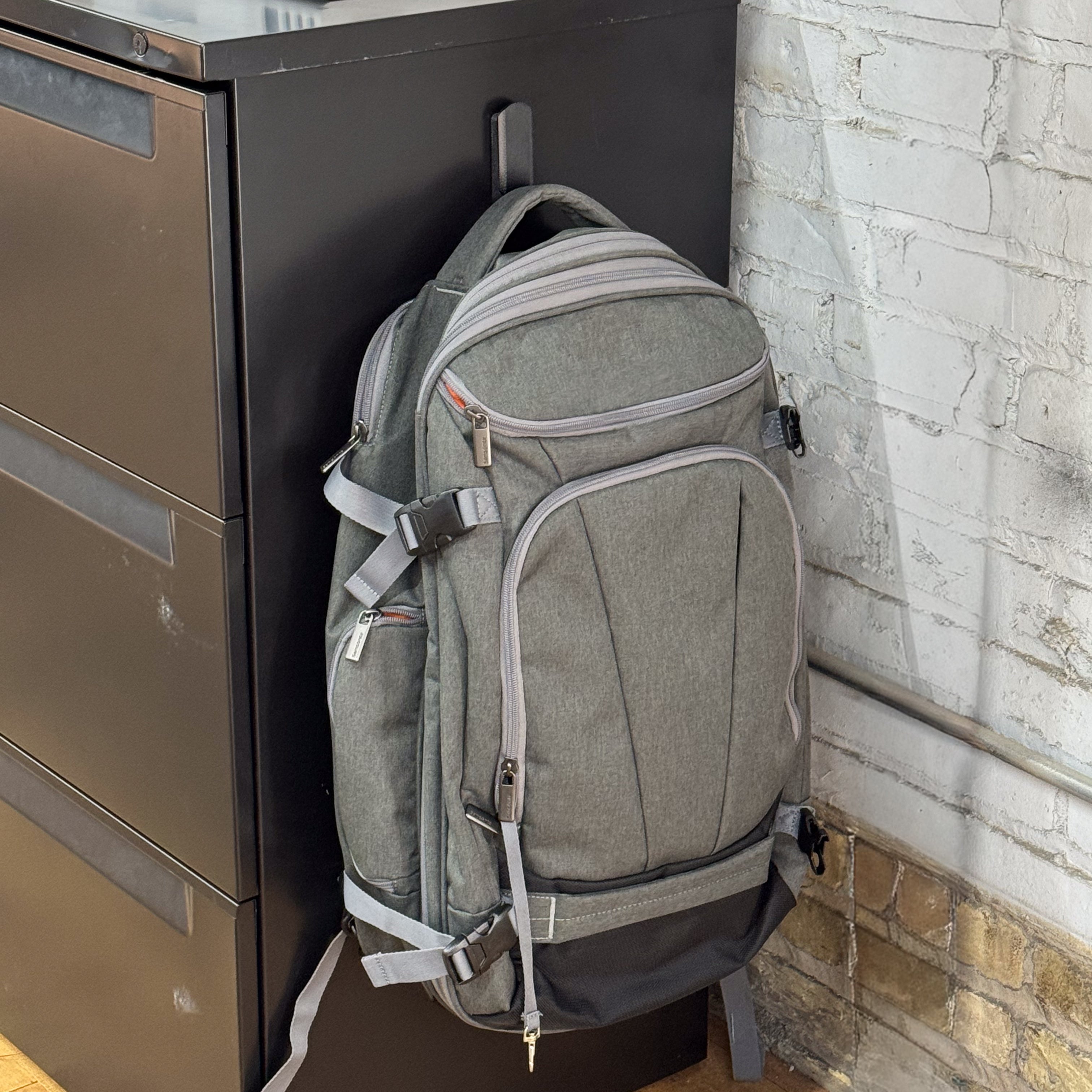 A gray backpack is hanging on a red Gator Magnetics hook attached to a flat, black steel filing cabinet in an indoor workspace corner.