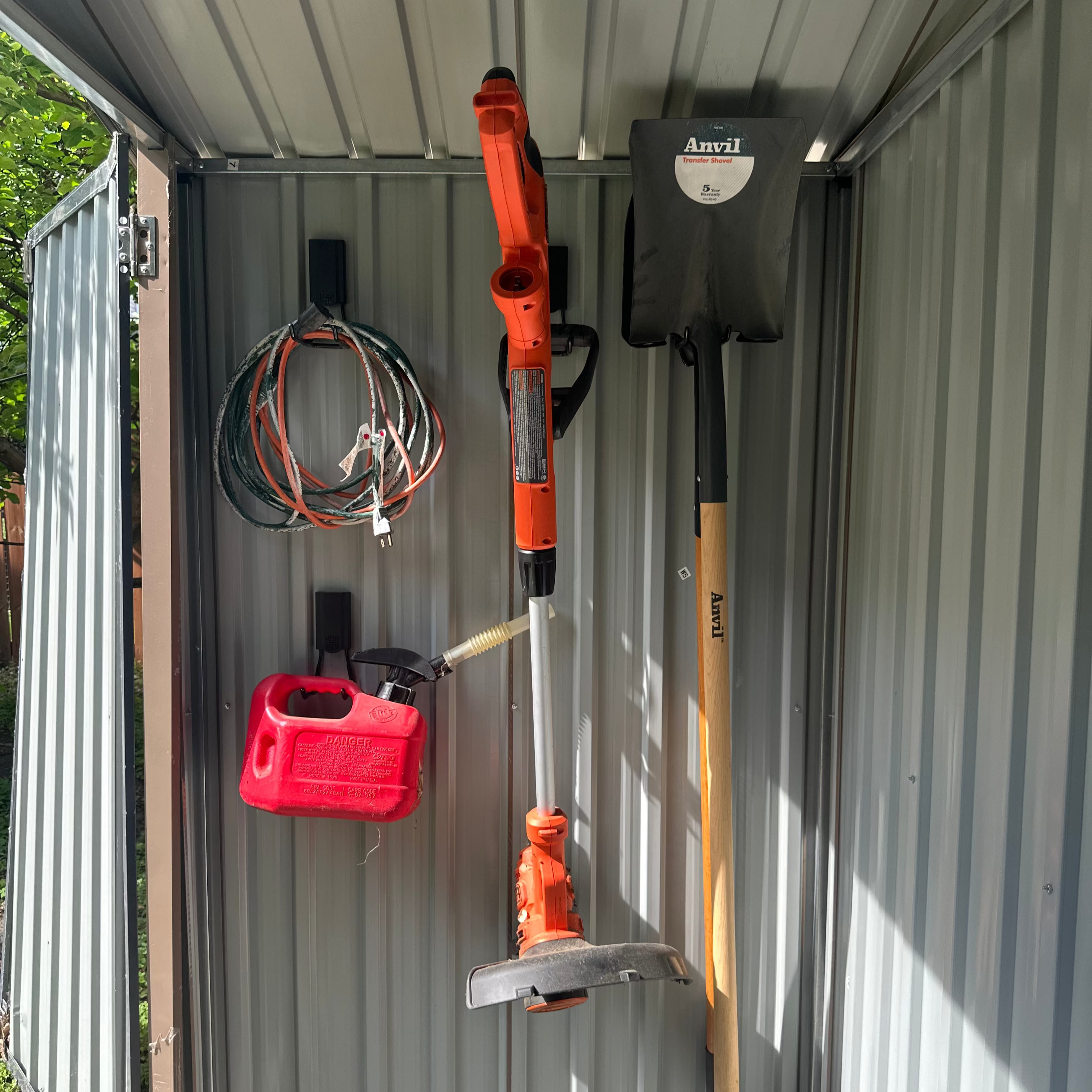 Red Gator Magnetics hooks hold an extension cord, red gasoline container, orange electric weed trimmer, and Anvil transfer shovel on the flat steel wall inside a small galvanized metal storage shed, demonstrating versatile garage storage for golf clubs and other equipment.
