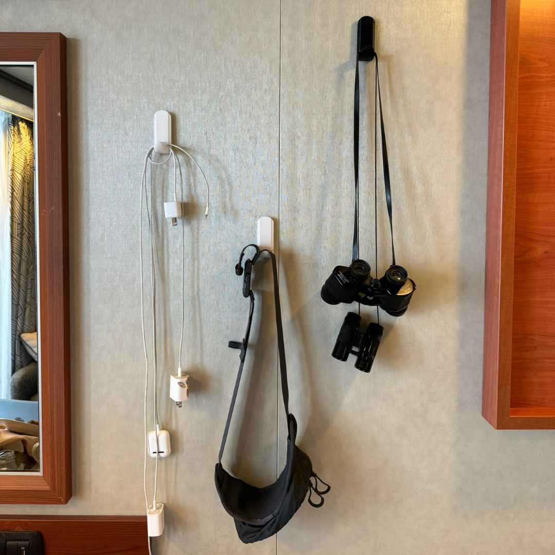 Three white hooks for hanging tools are attached to a steel wall panel, holding charging cables and adapters on the left hook, a black face mask on the middle hook, and two pairs of binoculars by their straps on the right hook.