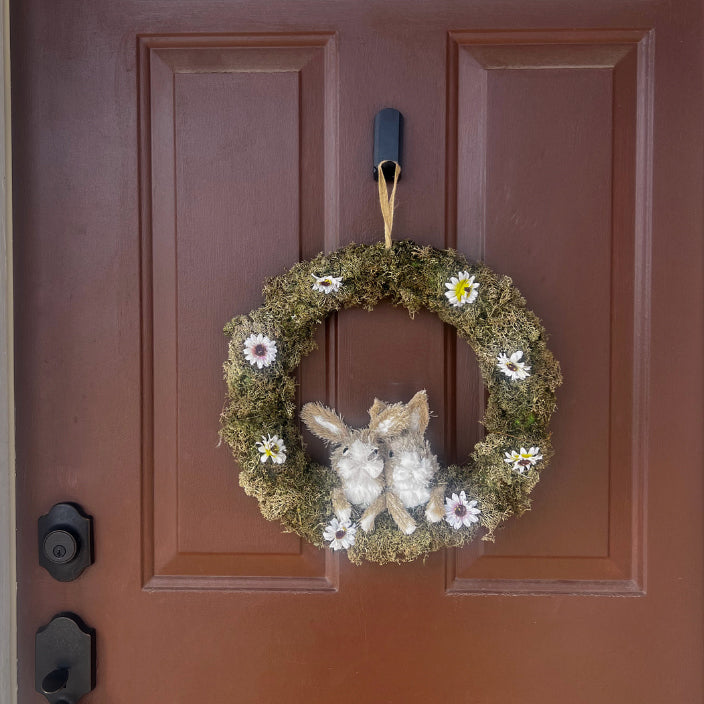 A decorative wreath made of moss and small white flowers is hung on a brown panel door using a black adhesive hook, illustrating a simple home application unrelated to Gator Magnetics but showing a common home backpack storage style for seasonal decor.