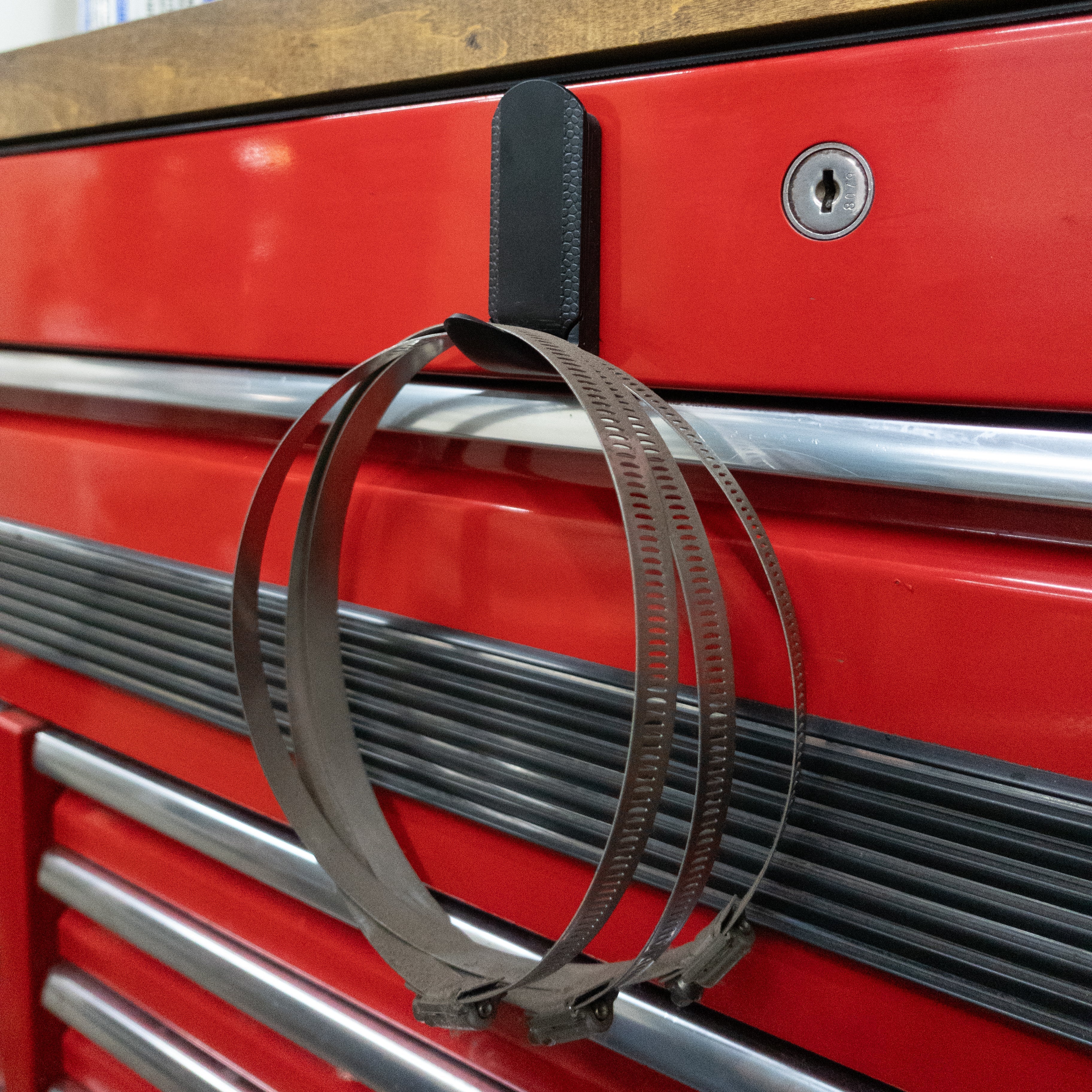 A black heavy duty utility hook from Gator Magnetics is magnetically attached to the front of a red steel toolbox, securely holding three large metal hose clamps.