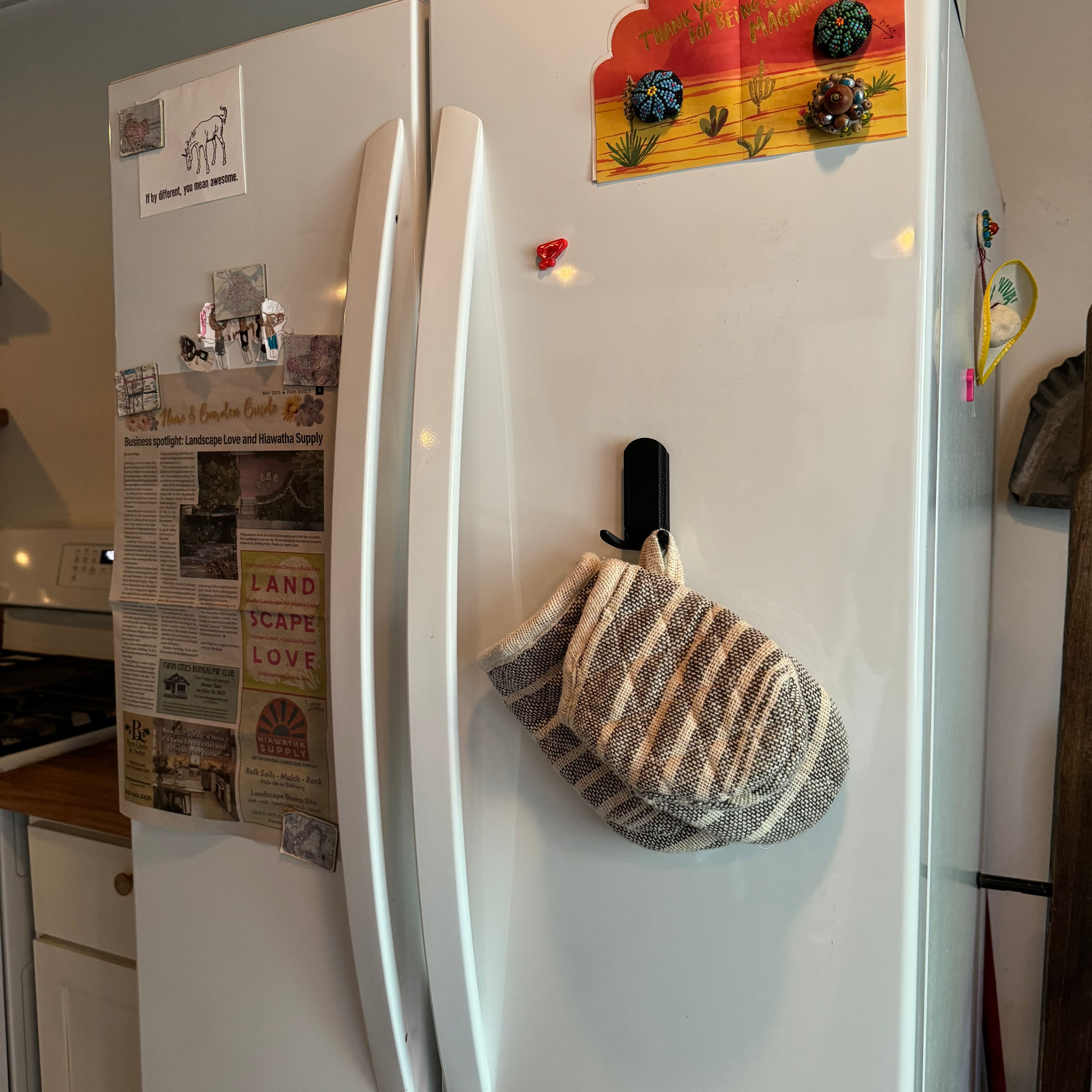 A black Gator Magnetics magnetic hook holds a striped oven mitt on the white steel surface of a refrigerator door in a kitchen, with various magnets and papers also attached nearby.