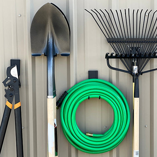 A steel pole barn wall features Gator Magnetics black magnetic hooks storing garden tools including a pair of loppers, a shovel, a green coiled garden hose, and a rake, demonstrating easy, secure storage for outdoor equipment.