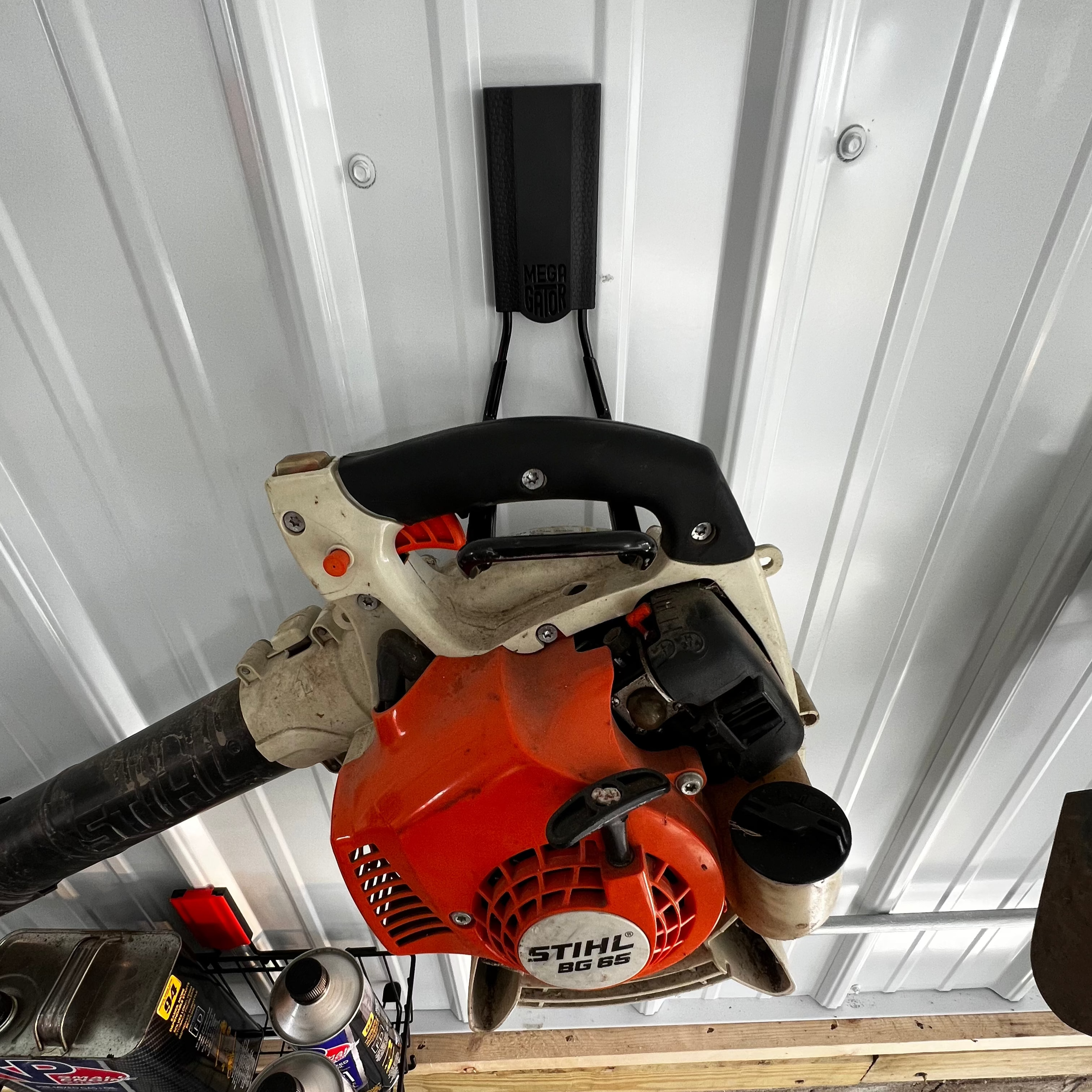 A Stihl BG 65 leaf blower is hung by its handle on a black Gator Magnetics hanger mounted to a white vertical steel wall panel, demonstrating a convenient leaf blower hanger for garage organization.
