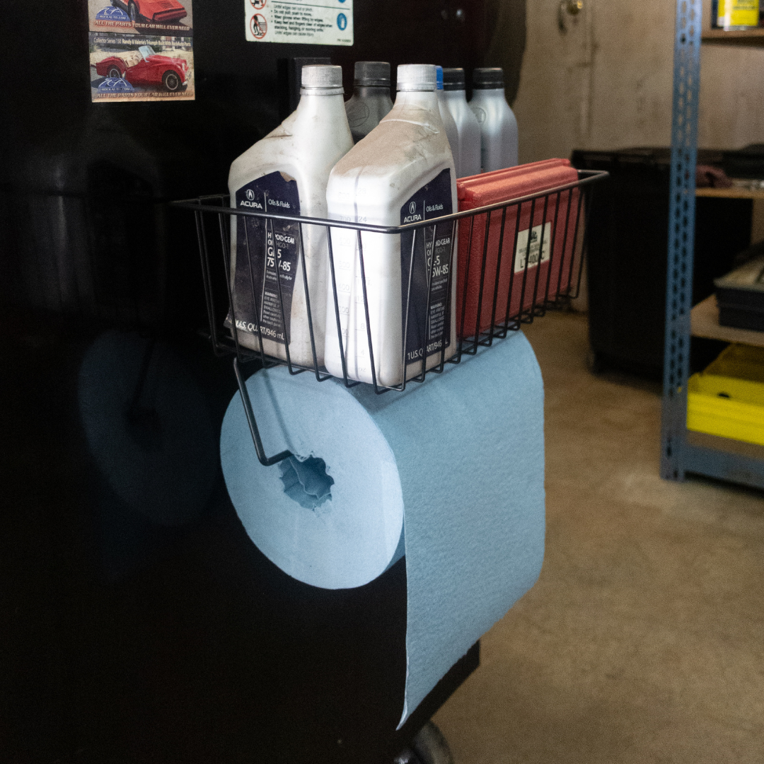 A small black basket from Gator Magnetics is magnetically mounted on a flat steel toolbox wall, storing several bottles of automotive fluids and a red box, with a roll of blue shop towels hanging beneath it.