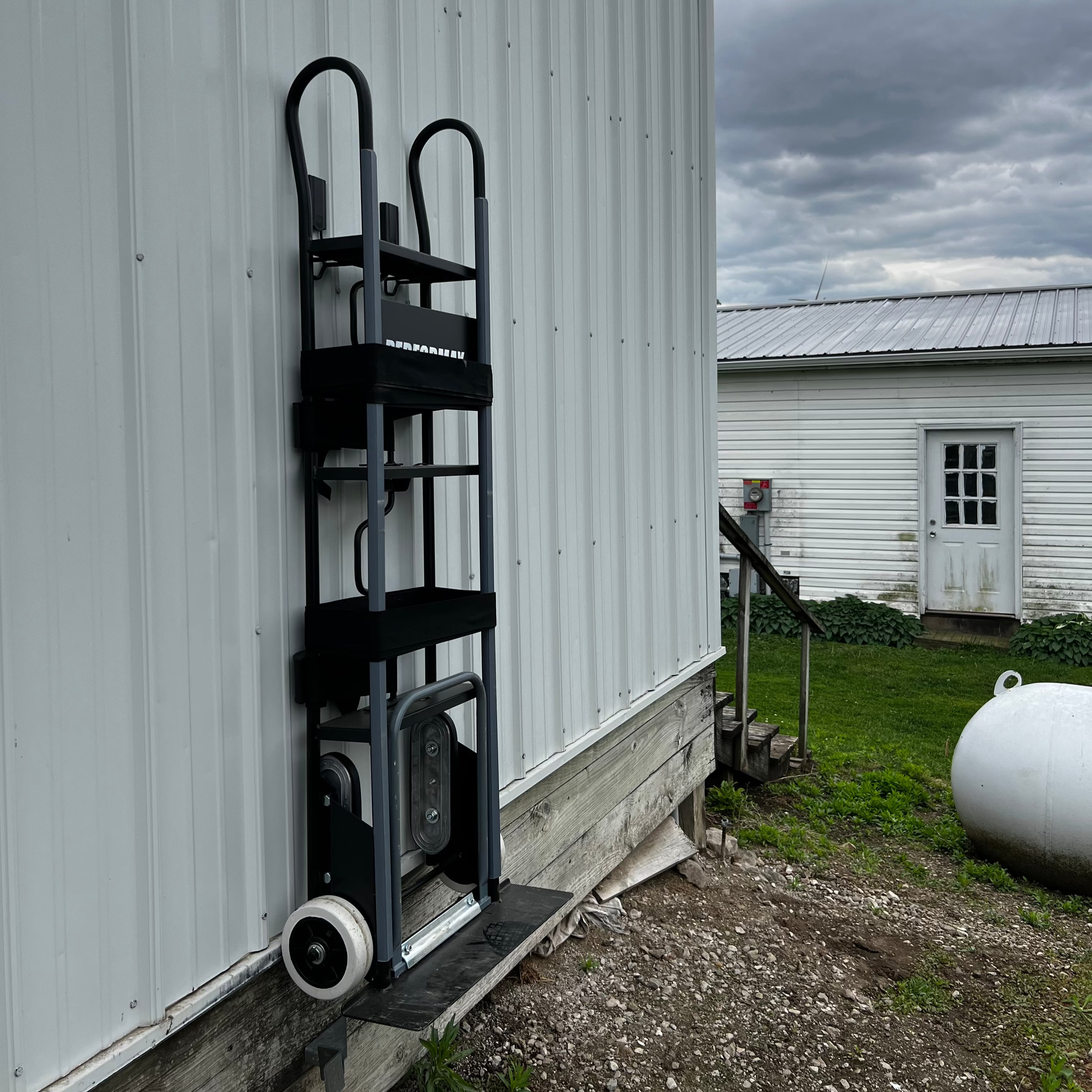 A hand truck is securely hung by its handle and base against a vertical corrugated steel wall panel of a pole barn exterior, demonstrating practical application of how to hang backpack blower or similar tools with Gator Magnetics products.