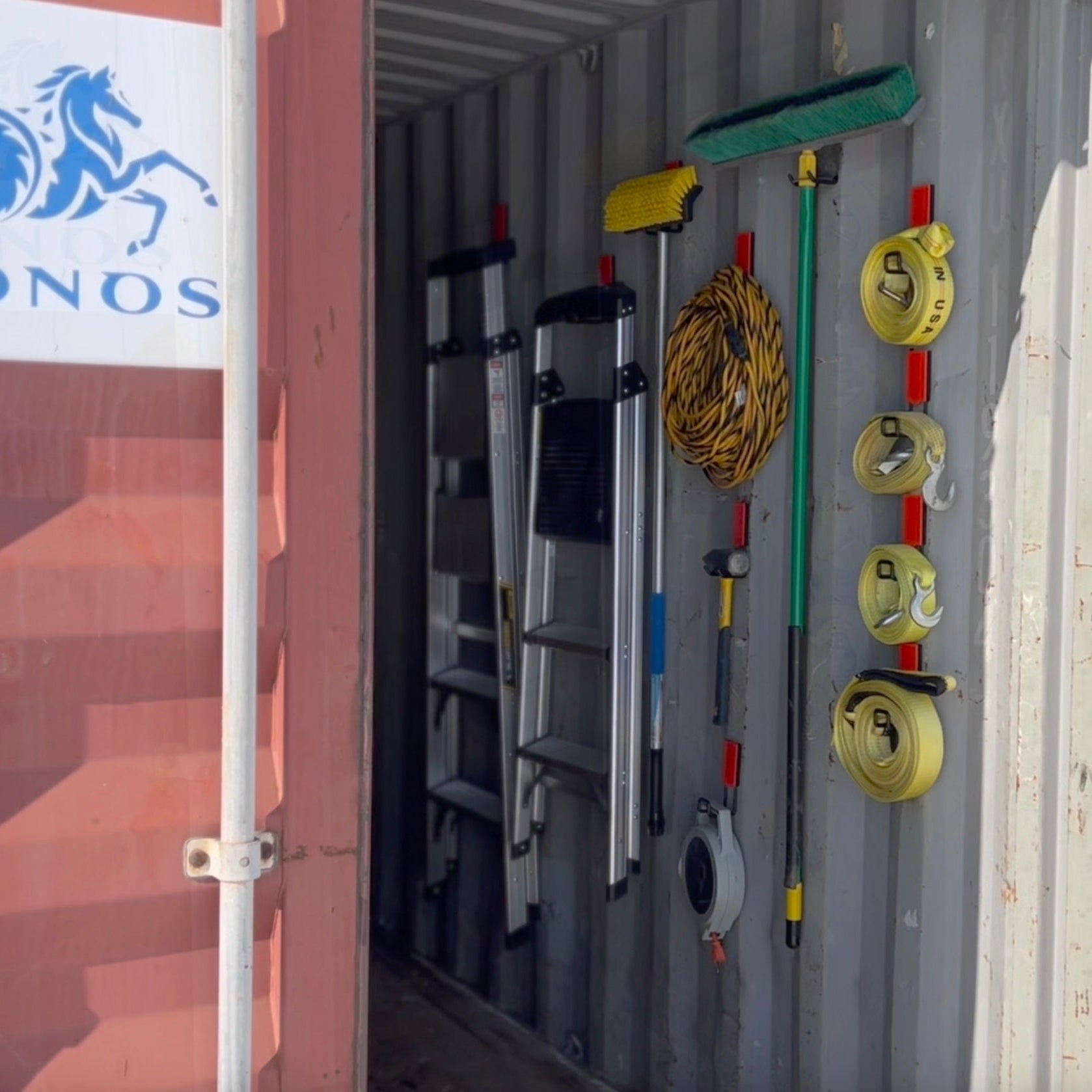 Two extension ladders, yellow extension cords, a broom, a hammer, a tape measure, and four yellow ratchet straps are hung on multiple red Gator Magnetics magnetic hooks mounted on the vertical steel wall inside a shipping container.