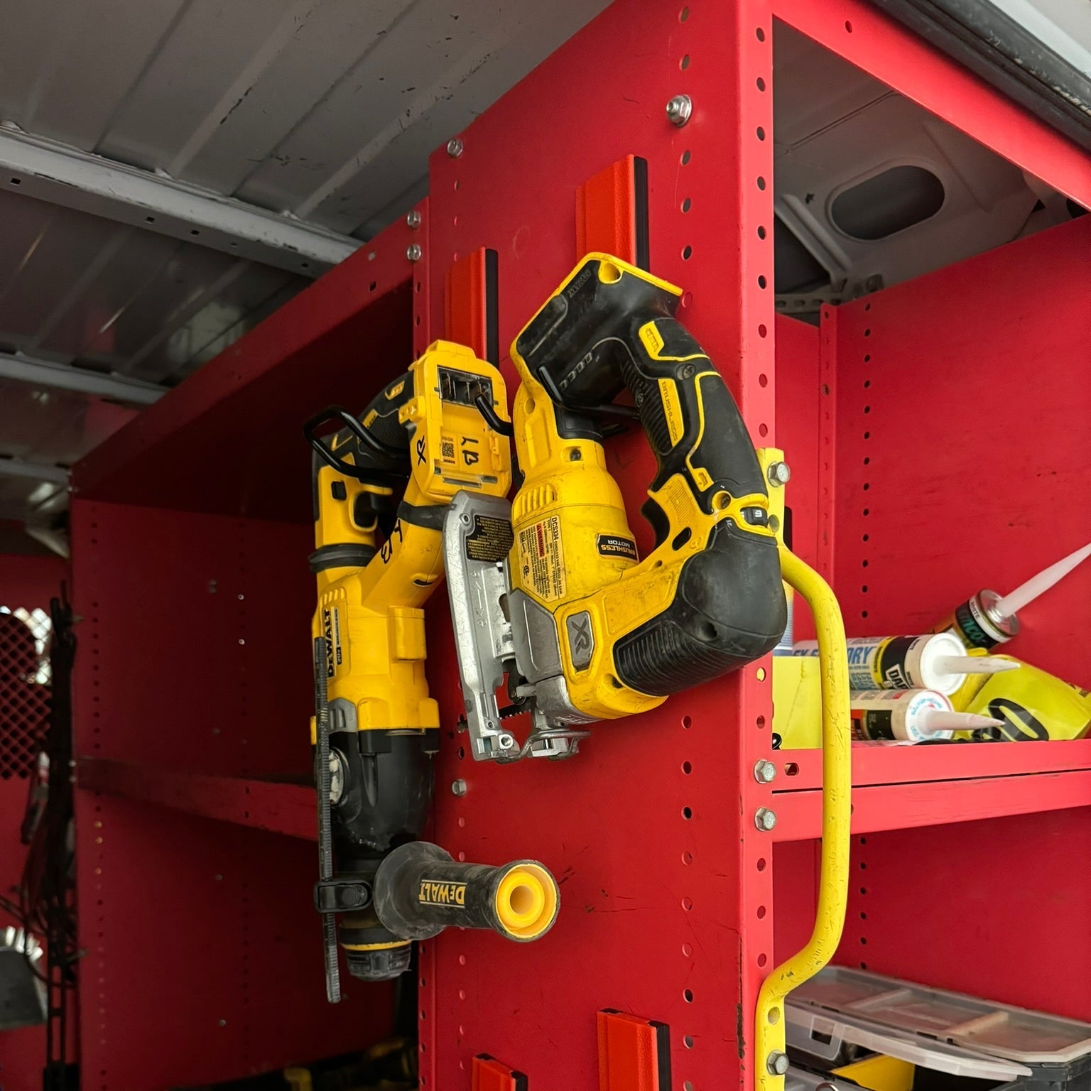 Two yellow power tools including a cordless jigsaw and reciprocal saw are securely hung by red open hooks on a red steel shelving unit inside a workshop van, showcasing practical tool storage without drilling or permanent hardware.