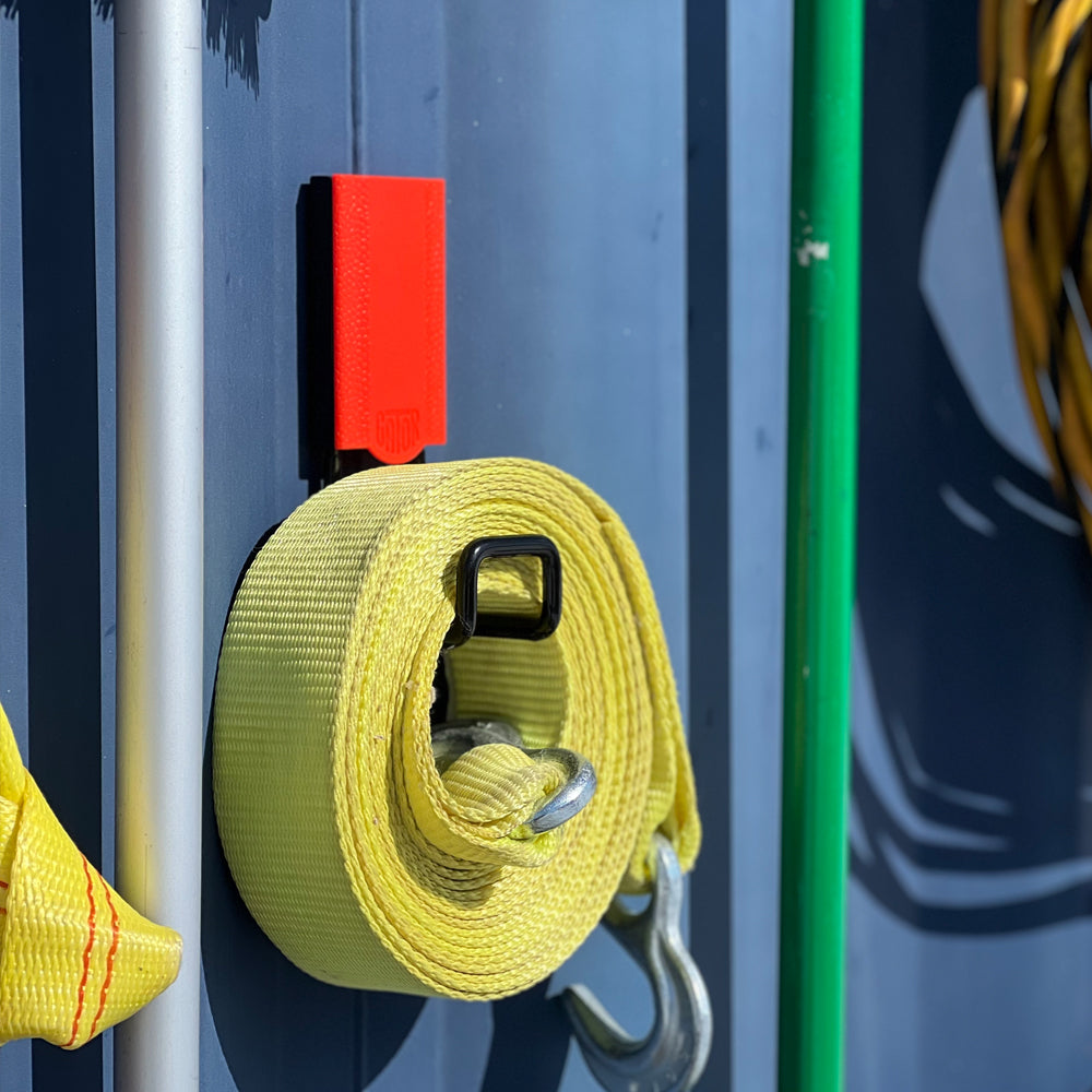 A yellow rolled-up ratchet tie-down strap with a metal hook hangs on a red Gator Magnetics magnetic hook attached to a flat steel shipping container wall panel, showcasing an efficient magnetic tool wall storage solution.