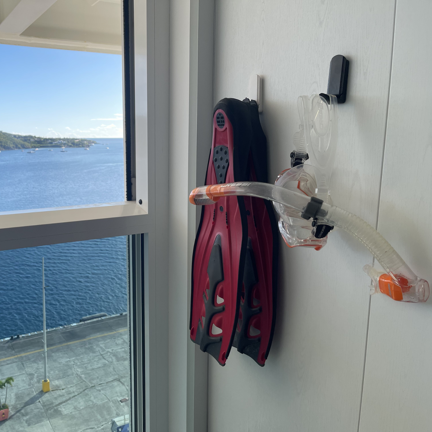 A pair of red and black swim fins and a snorkel mask with an attached snorkel tube are hanging on black MAGNETIC hooks mounted on a white painted steel wall panel near a window overlooking a waterfront.