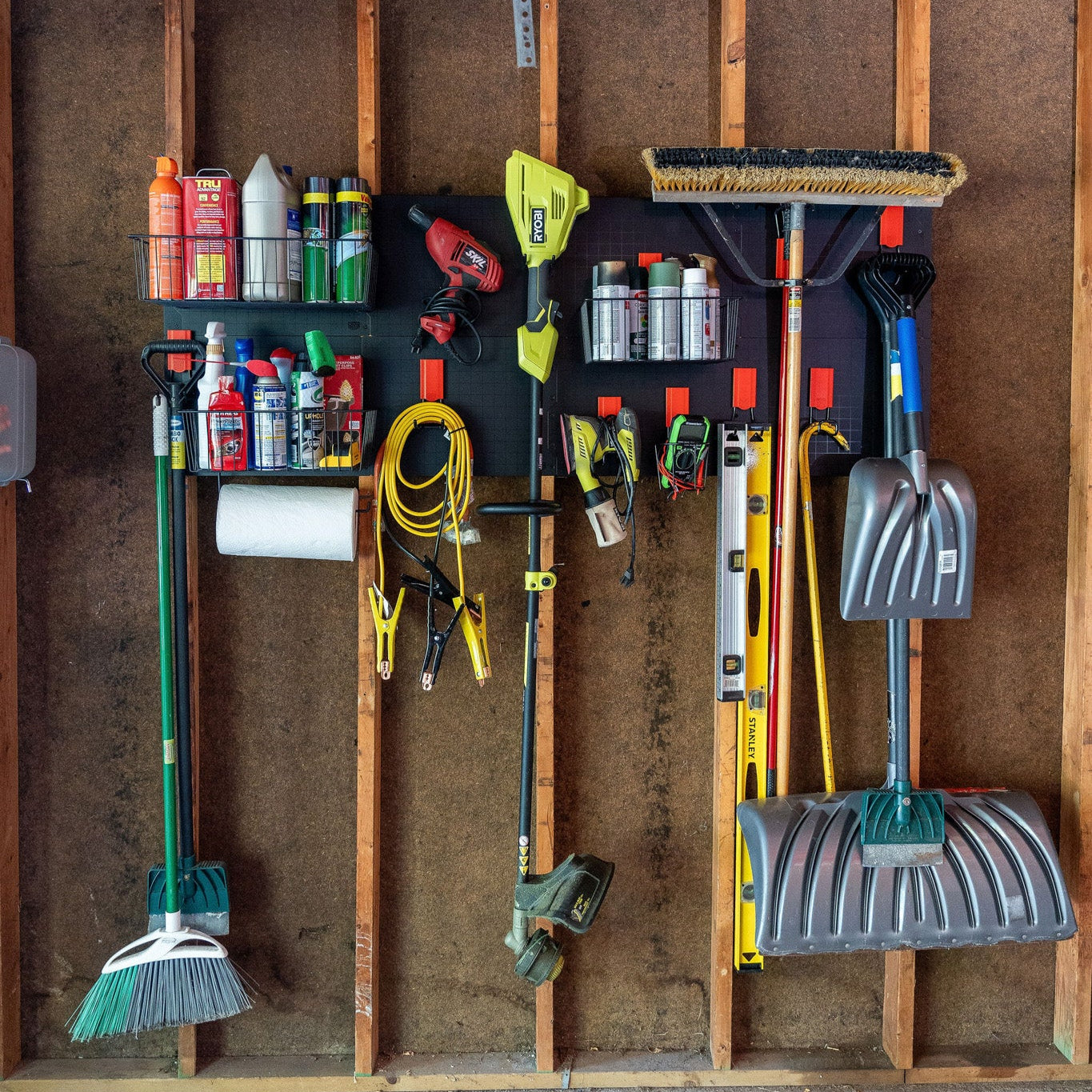 A garage wall storage hooks setup with Gator Steel Wall Panels mounted on vertical wooden studs, featuring magnetic storage baskets holding spray cans and fluids, red magnetic hooks suspending a broom, paper towel roll, jumper cables, electric weed trimmer, power drill, hand tools, a level, a rake, and multiple snow shovels inside a pole barn wall.