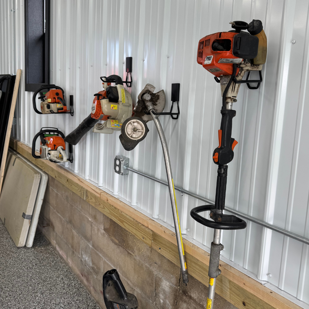 Four red industrial hooks from Gator Magnetics secure garden equipment including two orange and white gas-powered chainsaws, a leaf blower, and a string trimmer on a corrugated steel pole barn wall, demonstrating tool-free, damage-free mounting on a durable steel surface.
