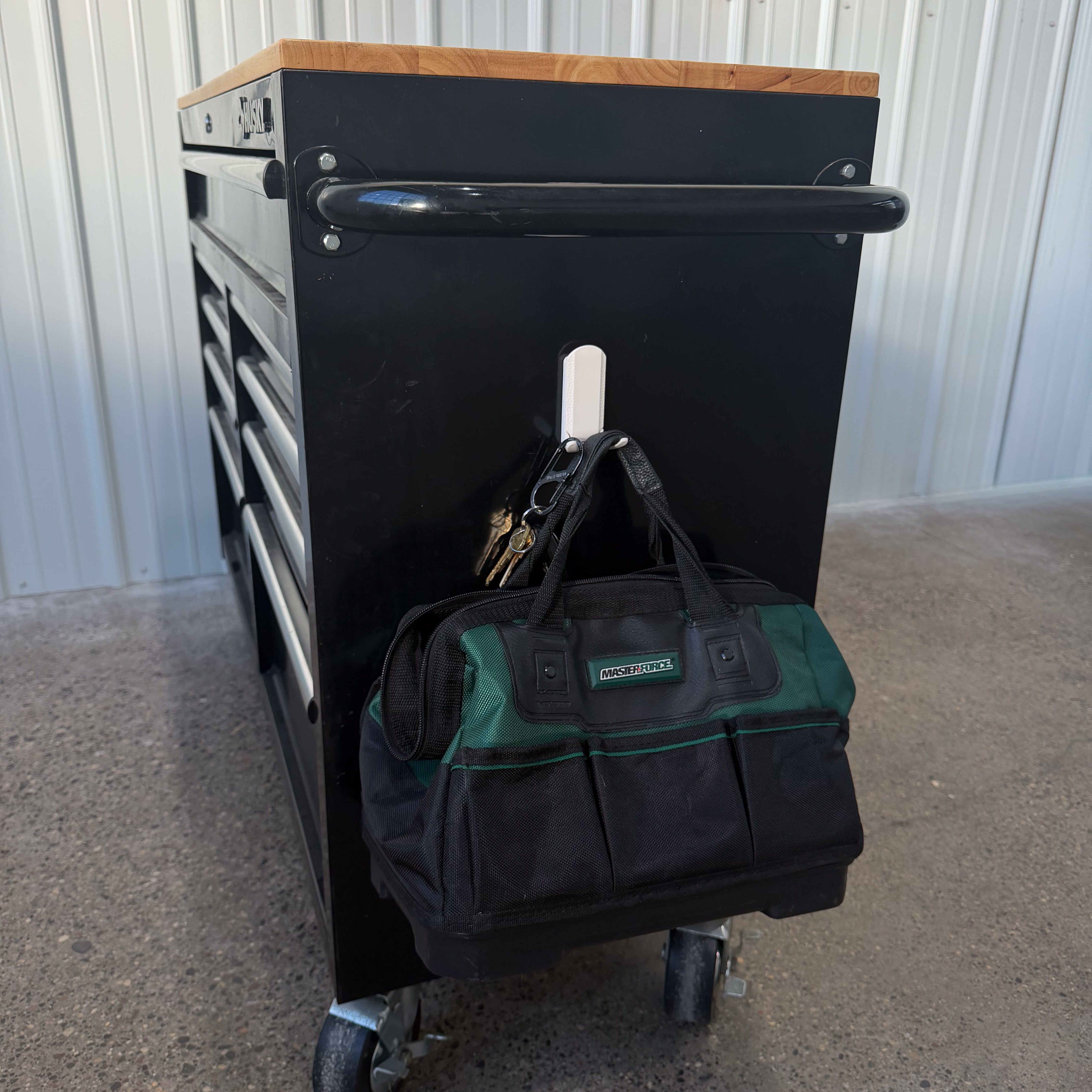 A white Gator Magnetic Hook with patented Easy-Off™ lever is magnetically holding a green and black tool bag and a set of keys on the black steel side panel of a rolling tool chest in a concrete floor workshop, demonstrating strong magnetic hold without permanent installation.