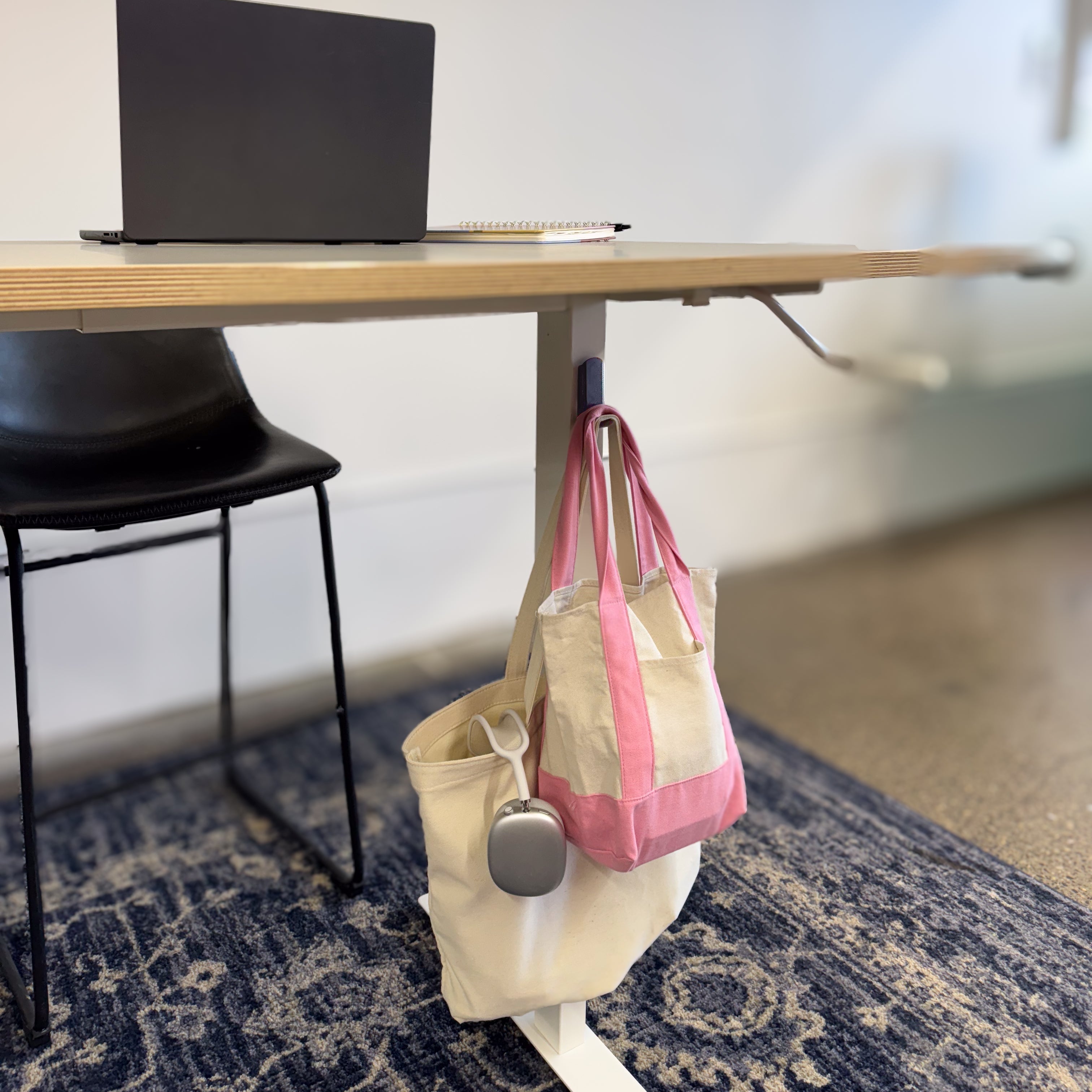 A Gator Magnetics magnetic hook holds two canvas tote bags, one with a yeti cup holder adapter attached, on the steel leg of a modern desk in an office setting with a laptop and notebook on the desk surface.