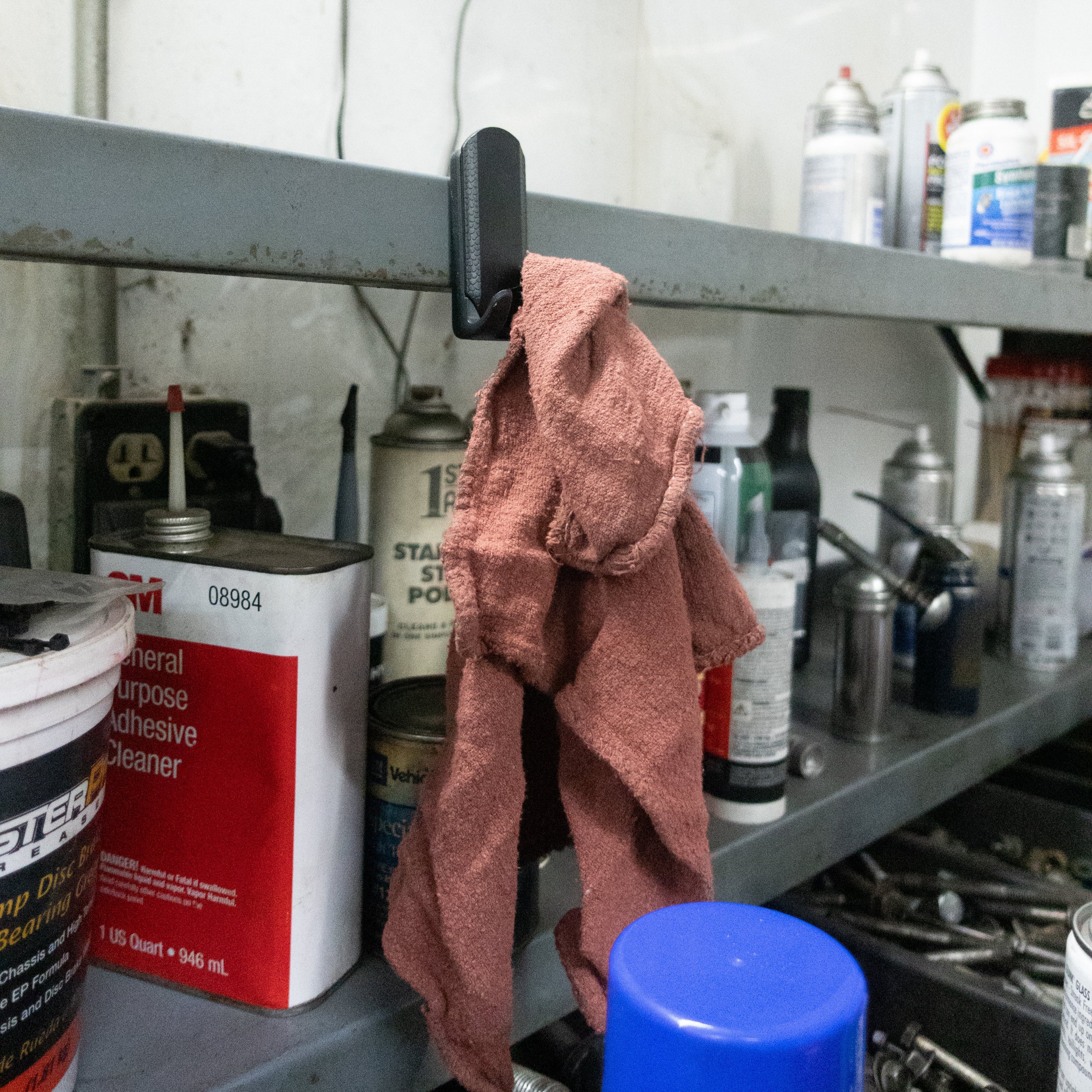 A black Gator Magnetics magnetic hook is attached to a steel shelf edge in a workshop, hanging a worn red rag, with various cans, spray bottles, and tools stored on the shelf and workbench below.