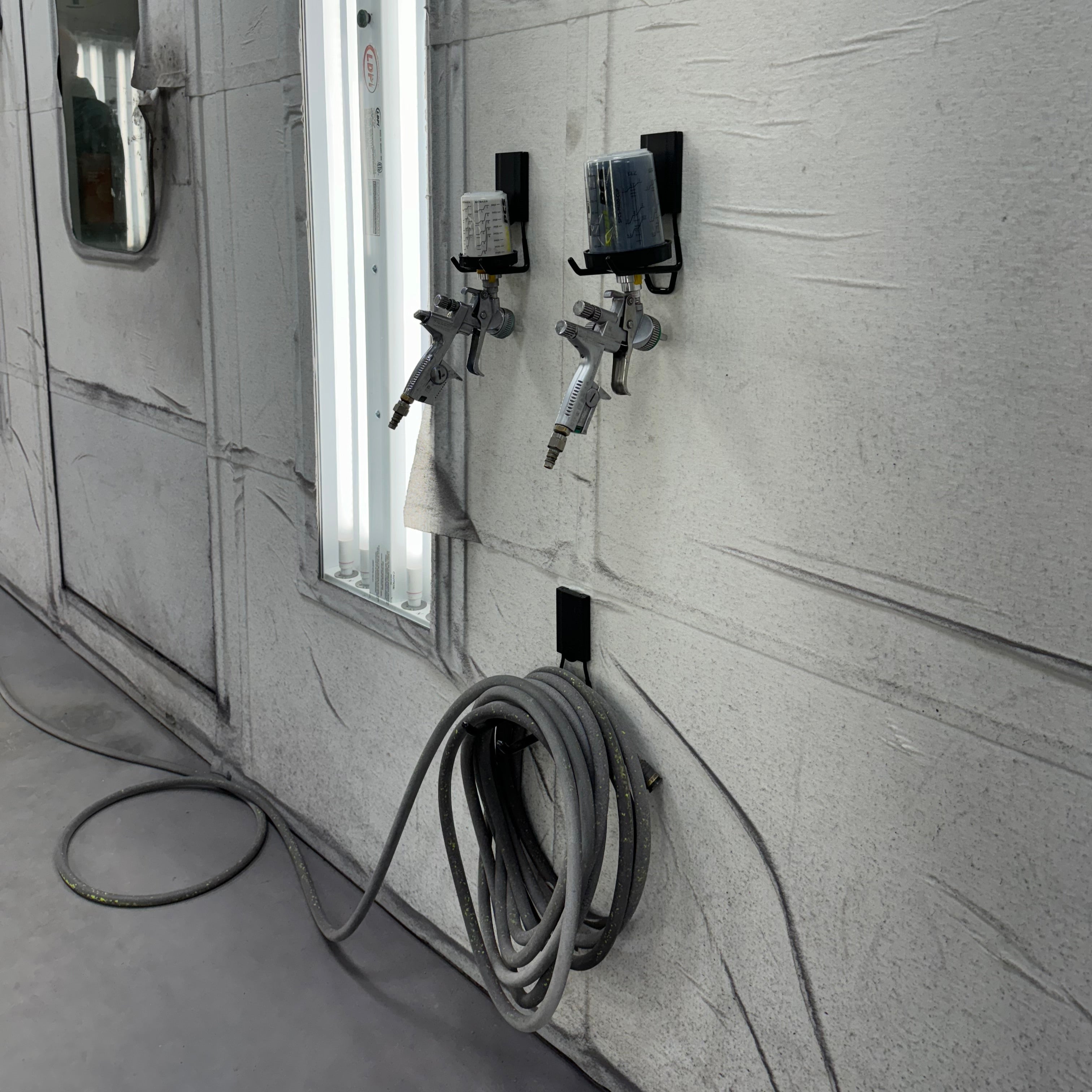 Two paint spray guns with attached mixing cups are held on black magnetic hooks alongside a coiled air hose stored on another black magnetic hook on a flat  steel wall, demonstrating a clean, tool-free organization approach useful for cruise storage hacks.