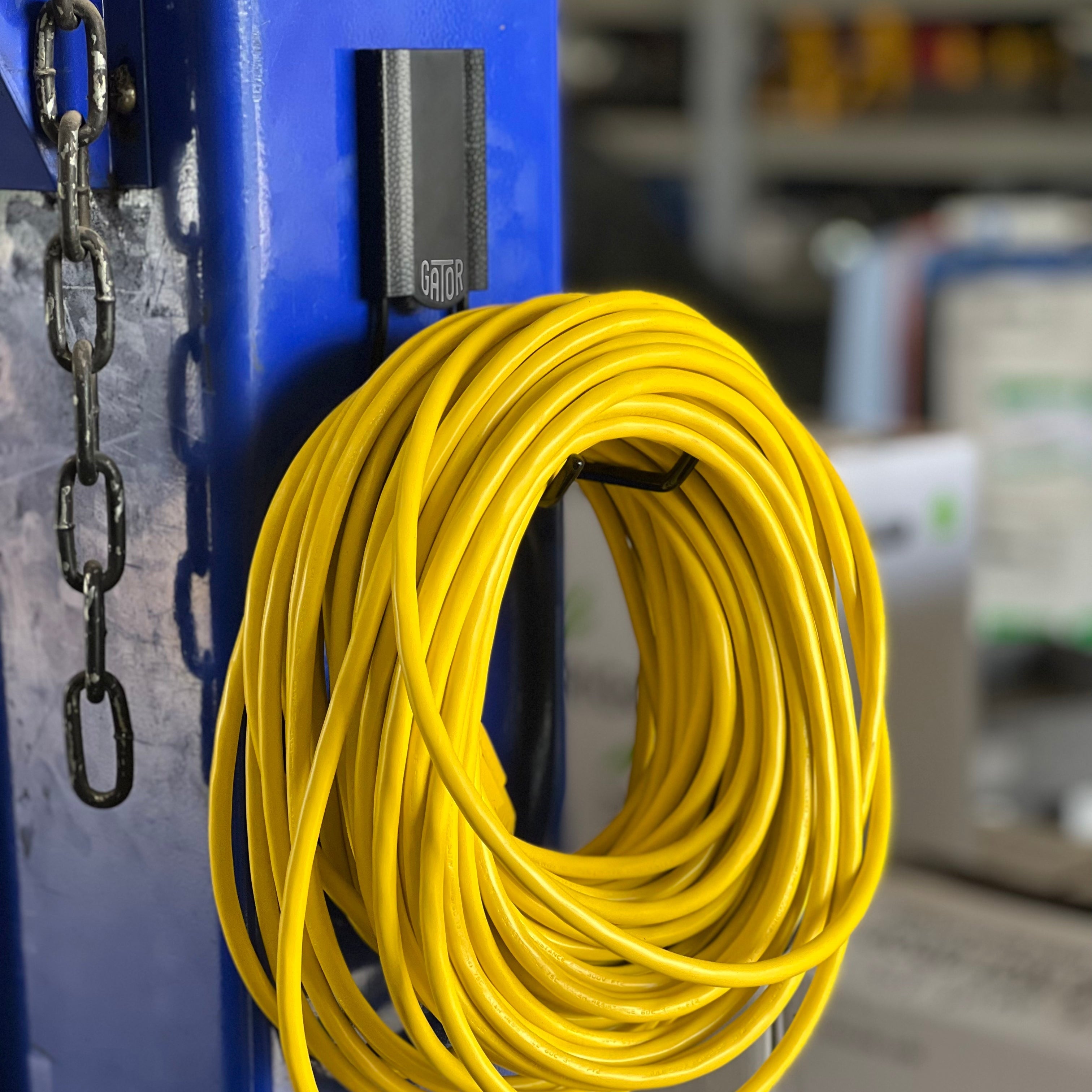 A bright yellow extension cord is neatly coiled and hung on a Gator Magnetics magnetic hook attached to a flat blue steel post in an industrial or workshop setting, demonstrating an effective way to organize backpack and cord storage without tools or permanent installation.