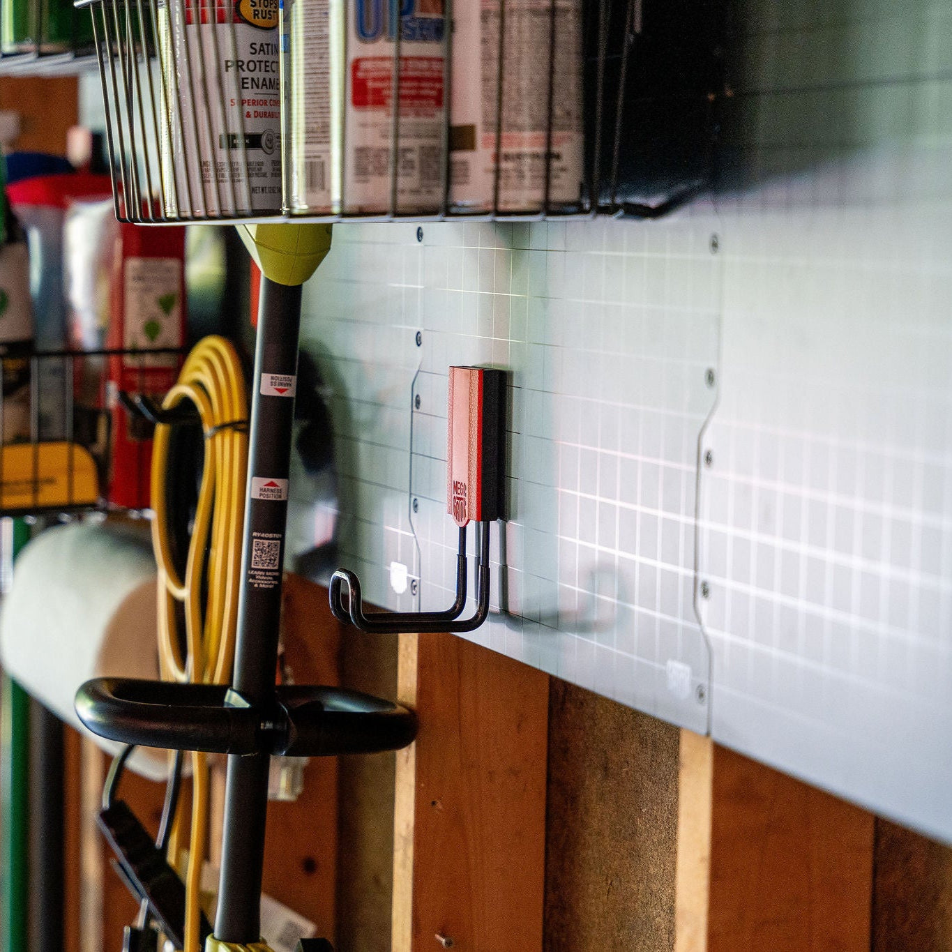 Two red magnetic hooks from Gator Magnetics are attached to a silver steel wall panel in a workshop, holding a garden tool with a black handle and a yellow hose coiled beside it.