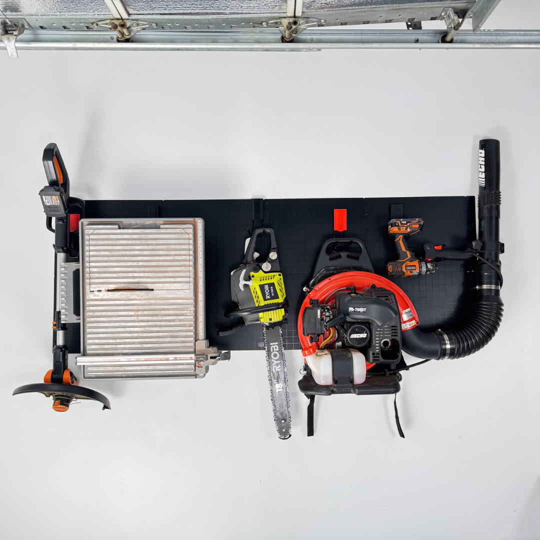 An array of garden tools including a string trimmer, a large metal tile cutter, a Ryobi chainsaw, an Echo backpack leaf blower, and a cordless drill are neatly hung on a black steel wall panel mounted inside a garage, demonstrating a magnetic hose hanger solution by Gator Magnetics for organized tool storage.