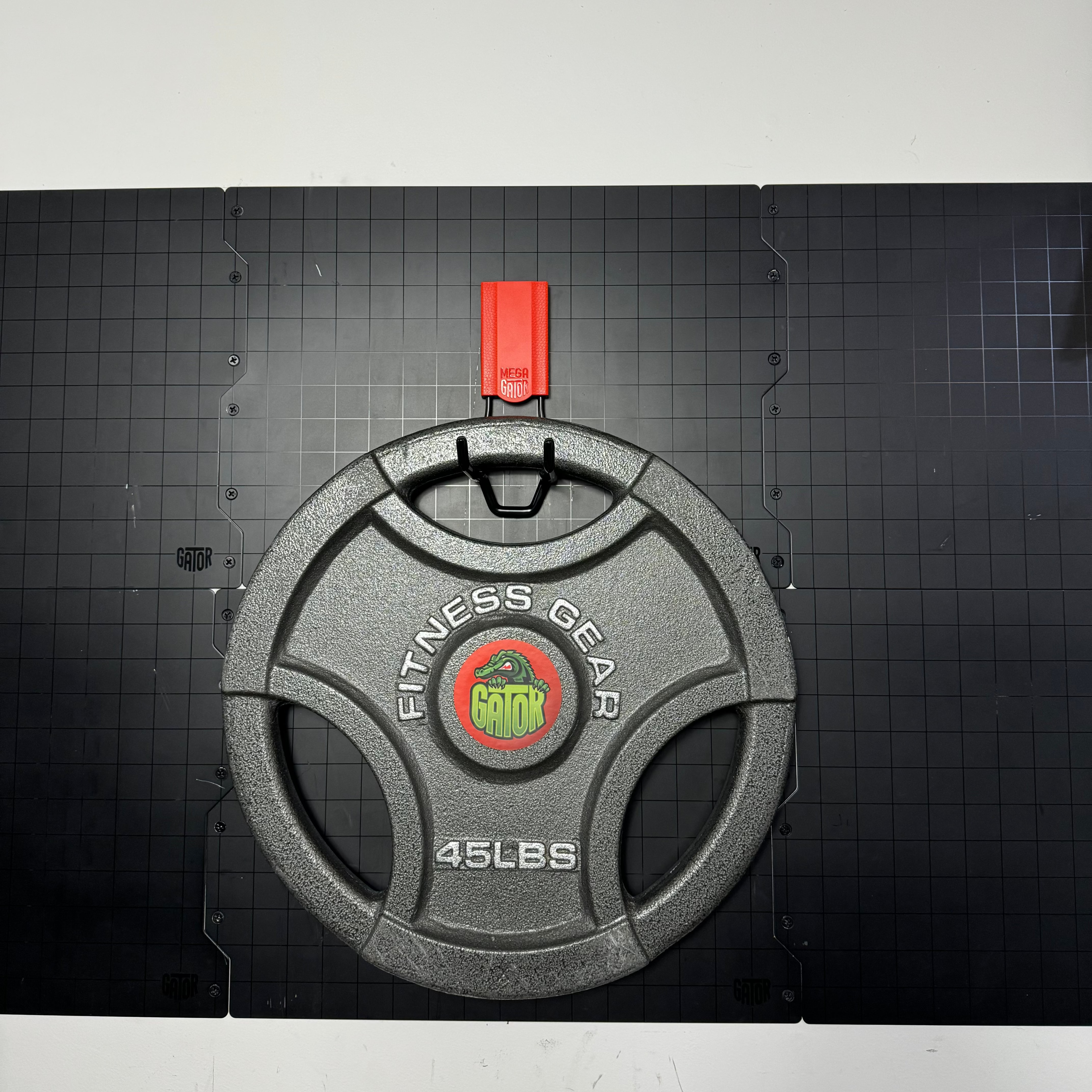 A 45 lb fitness gear weight plate is hanging from a red Gator Magnetics magnetic hook attached to a black Gator Steel Wall Panel mounted on a flat steel wall, demonstrating heavy-duty storage capacity ideal for gyms or workshops with a solid cup holder metal design.