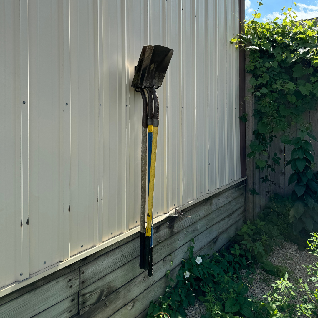 Two shovels with yellow and blue handles are hanging by red hooks on a vertical steel wall panel of a pole barn, demonstrating the use of hanging magnets for organized outdoor tool storage.