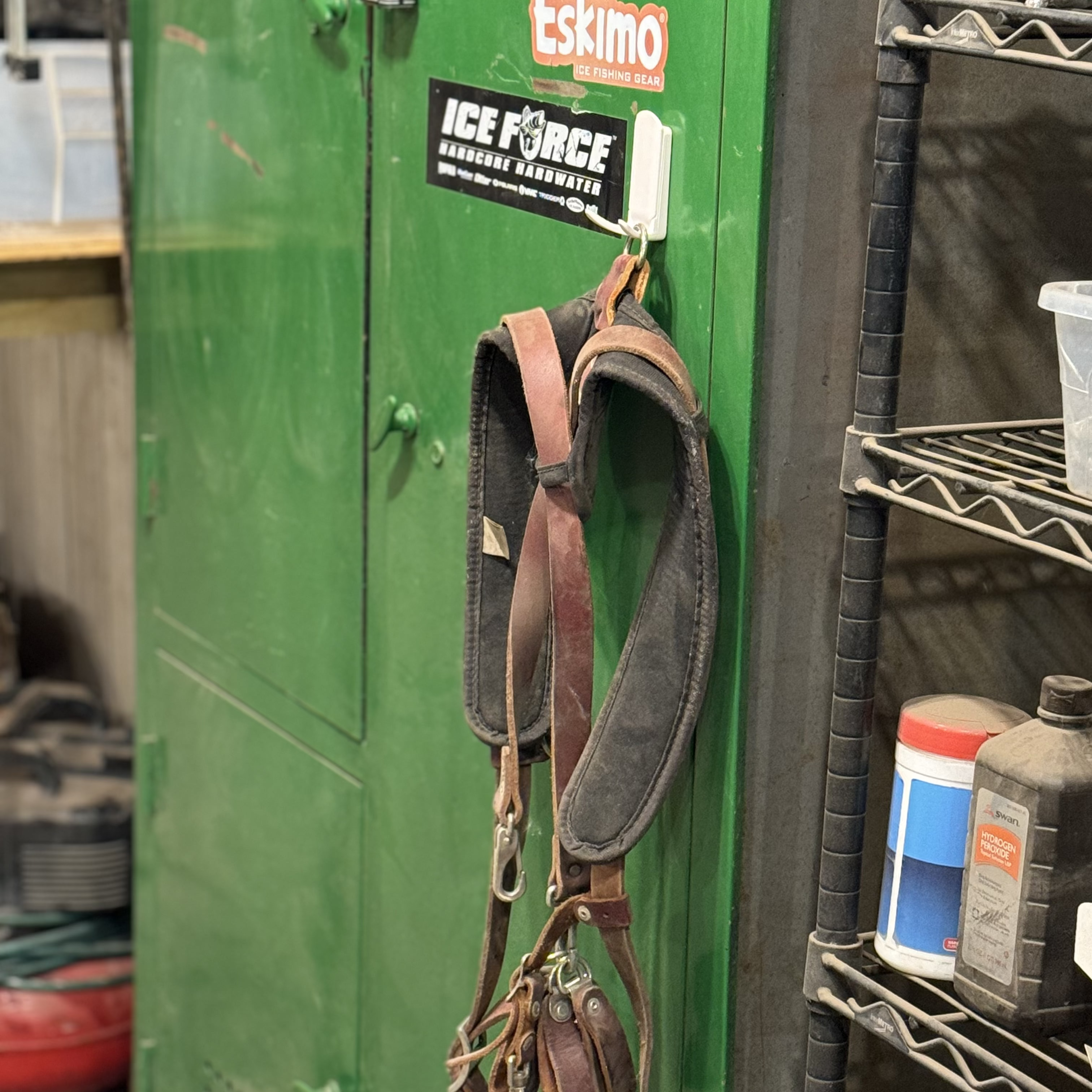 A brown leather harness is hanging from a white Gator Magnetics magnetic hook attached to a green  steel cabinet door in a workshop setting, with a black metal wire shelving unit holding containers nearby and a faint glimpse of a white small basket in the background.