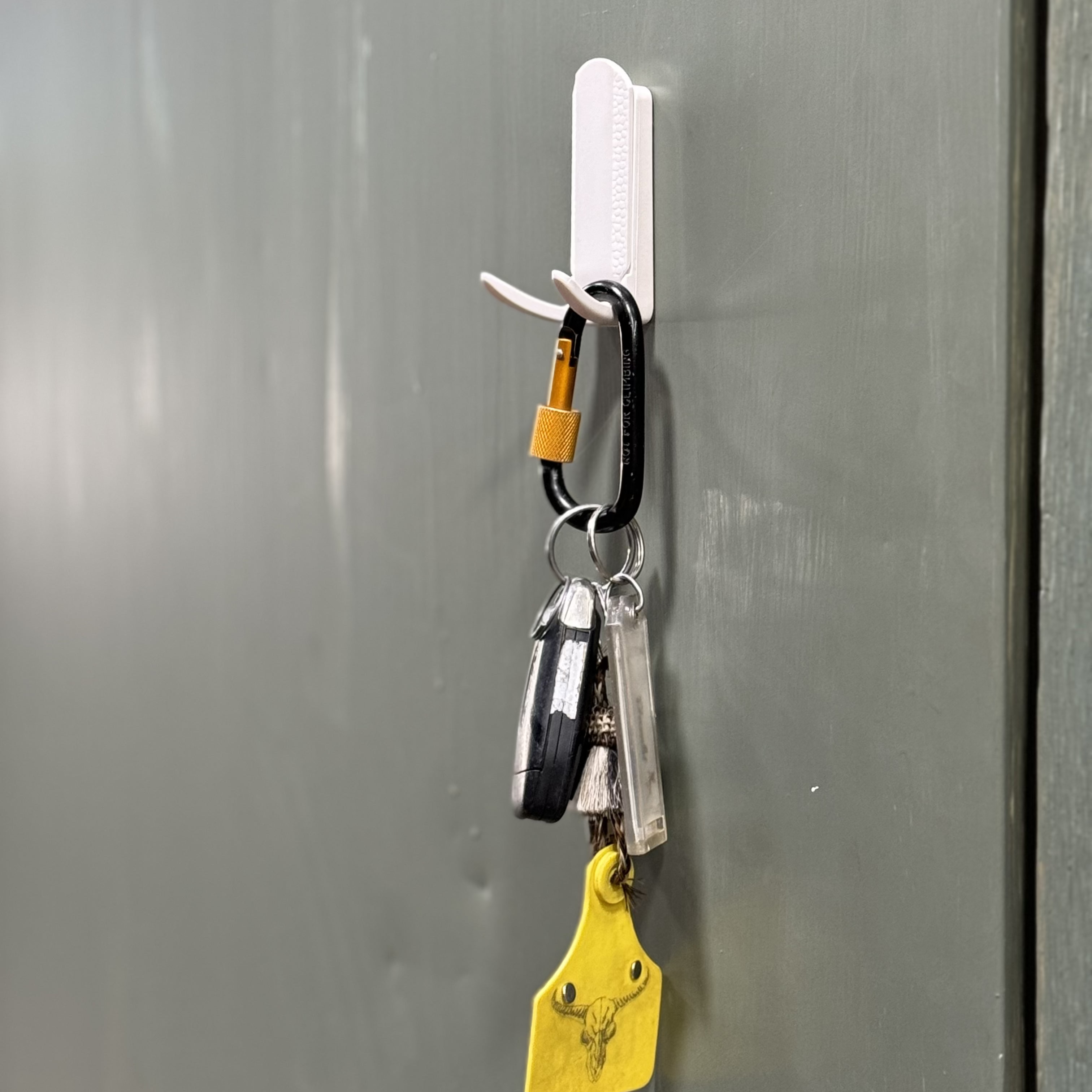 A white magnetic hook from Gator Magnetics is mounted tool-free on a flat steel wall panel, holding a black and orange carabiner with multiple keychains including a remote key fob, a clear plastic key tag, and a yellow leather key tag with a deer skull design, demonstrating practical golf organization for compact steel storage.